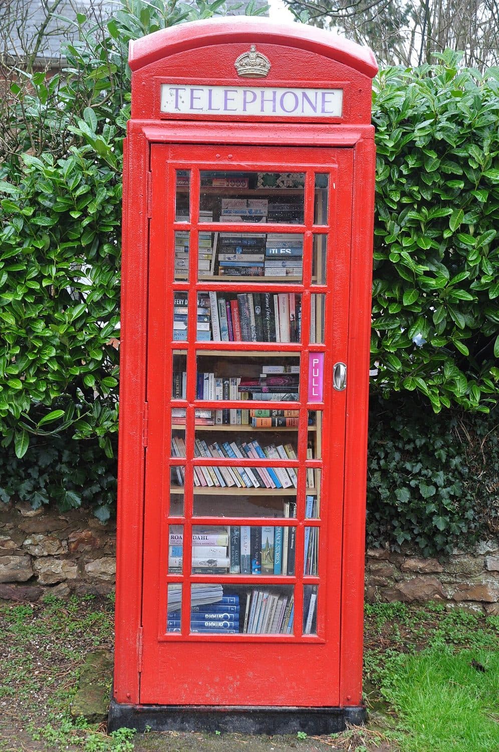 Una cabina telefonica rossa trasformata in biblioteca, tra una siepe e un muretto di pietra